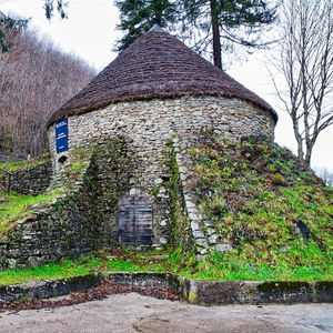A photograph of a rustic, circular stone building with a thatched roof, nestled amidst a lush green hillside. The building appears to be an ancient structure, possibly a granary or a well-preserved historical site. 